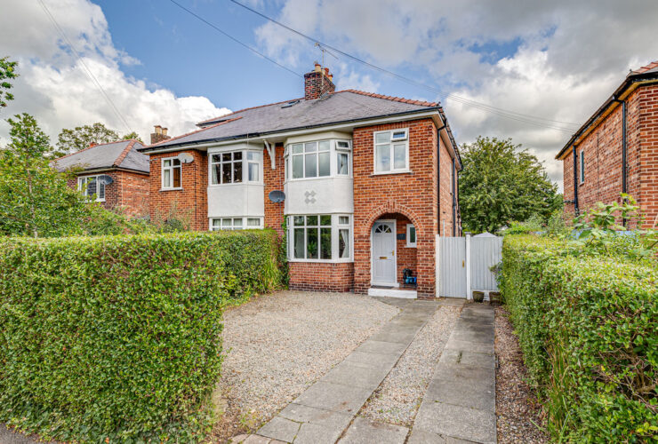A red brick semi detached house with bay windows. In the foreground a gravel driveway with a stone slab path leading to a light blue front door. The driveway boundary is marked with green hedges.